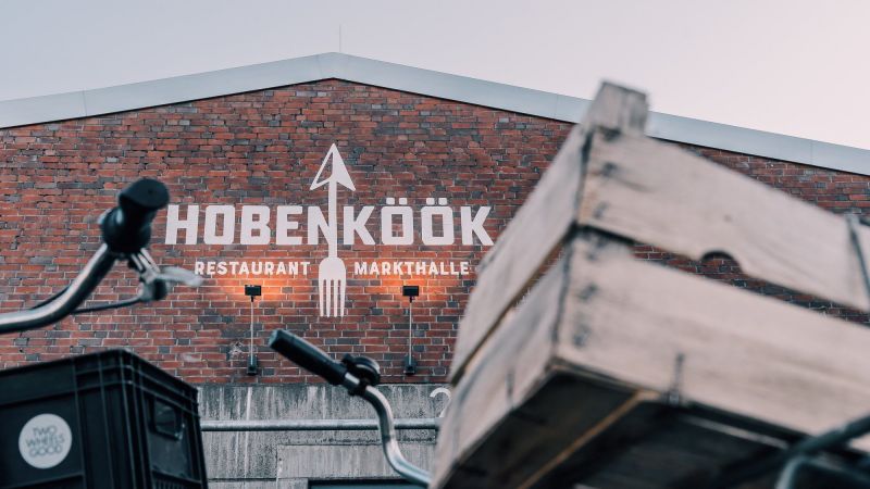 Außenansicht der HOBENKÖÖK-Fassade aus rotem Backstein mit Logo; Markthallen-Sicht, im Vordergrund Holzkisten und Wagen.