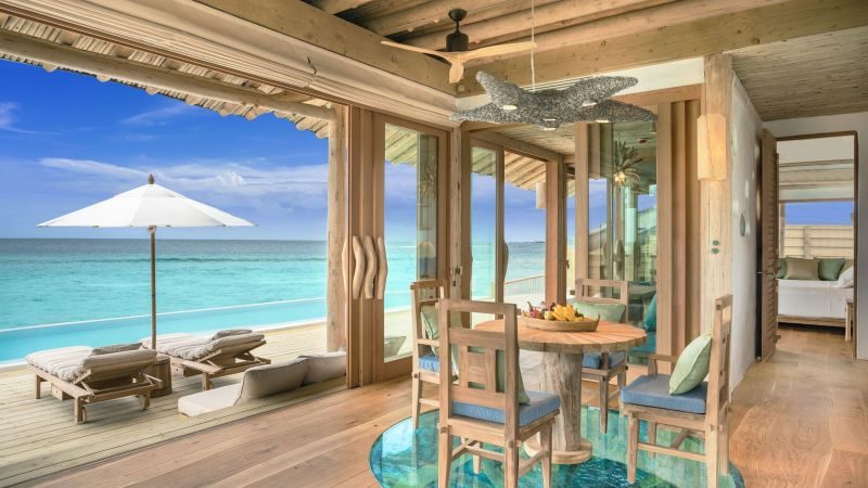 Wohnbereich einer Soneva Fushi Water Retreat mit Blick aufs Meer, Holzmöbel, Esstisch, Glastüren und Liegen mit Sonnenschirm.