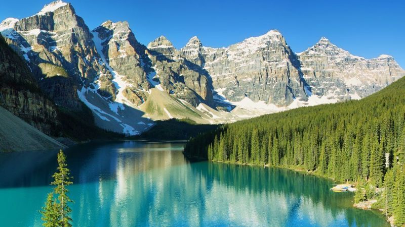 Türkisfarbener Bergsee, von dichten Nadelwäldern umgeben, schneebedeckte Berge im Hintergrund, Nordamerika