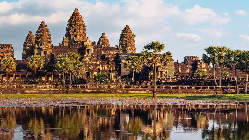 Angkor-Wat-Tempelkomplex am Wasser, Palmen im Vordergrund, Reflexion im See, goldenes Licht.