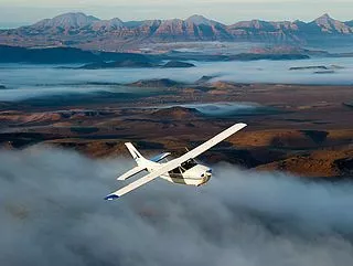 Weißes Kleinflugzeug fliegt über nebelige Namibia-Wüstenlandschaft mit Bergkette im Hintergrund.