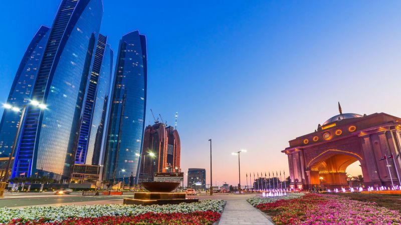 Abu Dhabi Skyline bei Dämmerung: blaue Glashochhäuser links und ein heller Torbogen rechts, Blumenbeete im Vordergrund