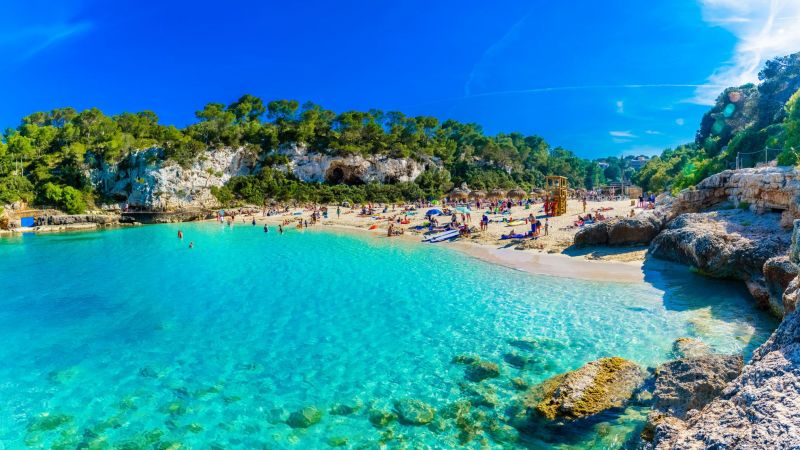 Panoramabild Cala Llombards Strand mit türkisblauem Wasser, sandigem Ufer, Felsen und Urlaubern, Balearen