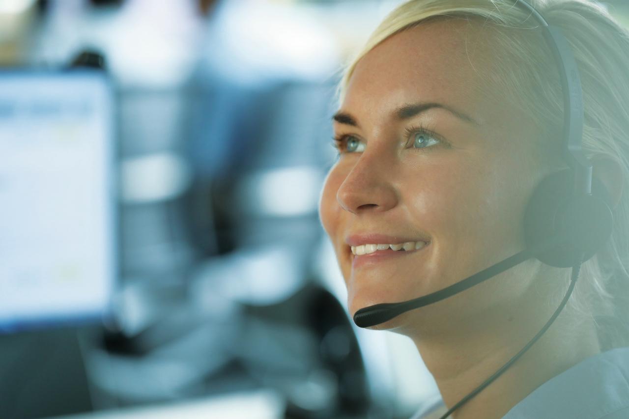 Frau mit Headset im Callcenter, lächelnd, seitlicher Blick, unscharfer Hintergrund.