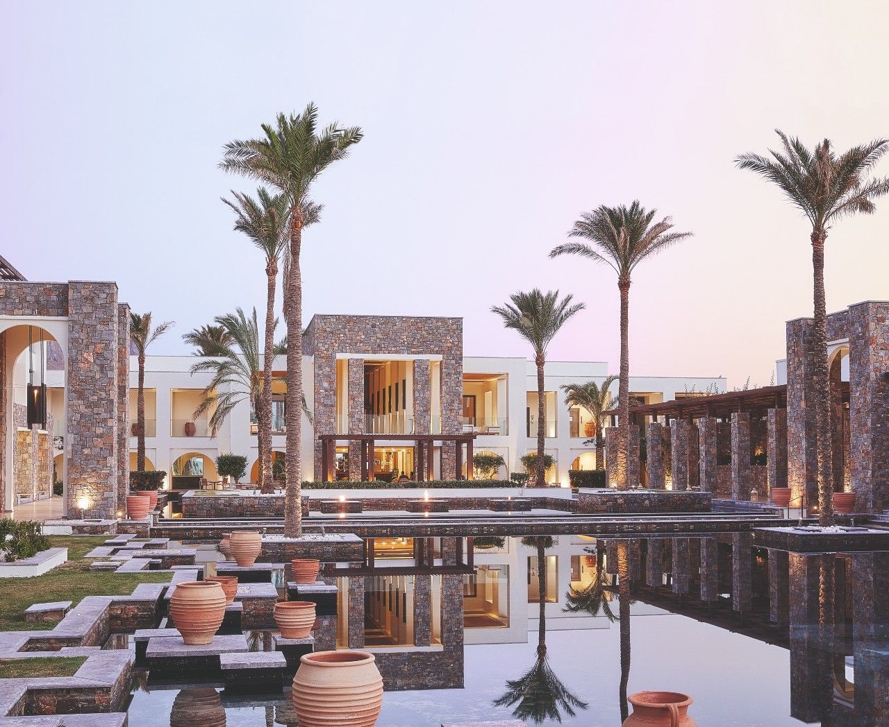 Außenansicht eines luxuriösen Resort-Komplexes mit Steinarchitektur, Palmen und spiegelndem Wasserbecken.