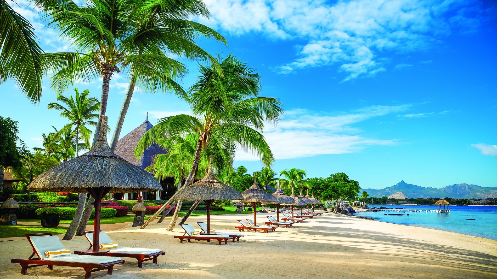 Weicher Sandstrand mit Palmen, mehreren Strohut-Schirmen und Liegen am klaren Meer vor Mauritius, Oberoi Resorts