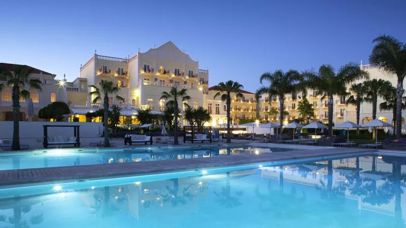 Außenansicht eines eleganten Domes Resorts-Komplexes am Abend, beleuchtete Fassade, Palmen, Pool mit Spiegelung.