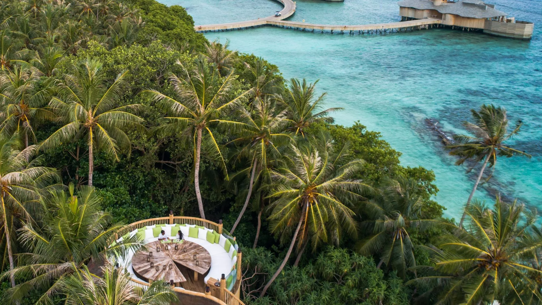 Luftaufnahme eines Malediven-Resorts: Palmen, über Wasser-Bungalows und eine runde Zipline-Dining-Plattform im Vordergrund.