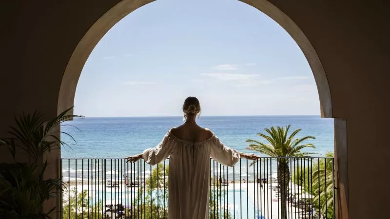 Frau in off-Shoulder-Kleid steht auf Balkon zum Meer, Domes Resorts; Geländer, Palmen und Poolbereich sichtbar.