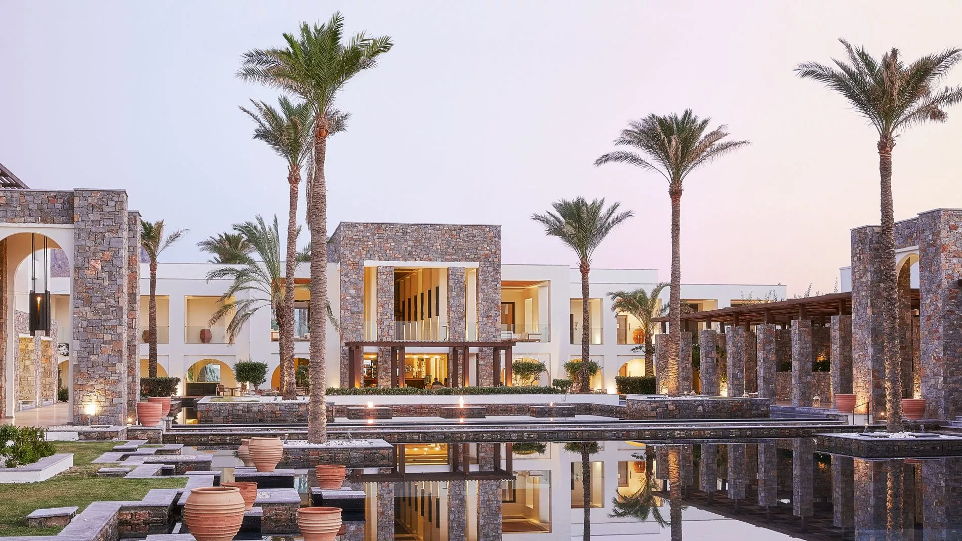 Grecotel Amirandes Innenhof mit Palmen, Steinarchitektur, Terrassen und reflektierenden Wasserbecken bei Abendlicht. Grecotel Amirandes Innenhof mit Palmen, Steinarchitektur, Terrassen und reflektierenden Wasserbecken bei Abendlicht.