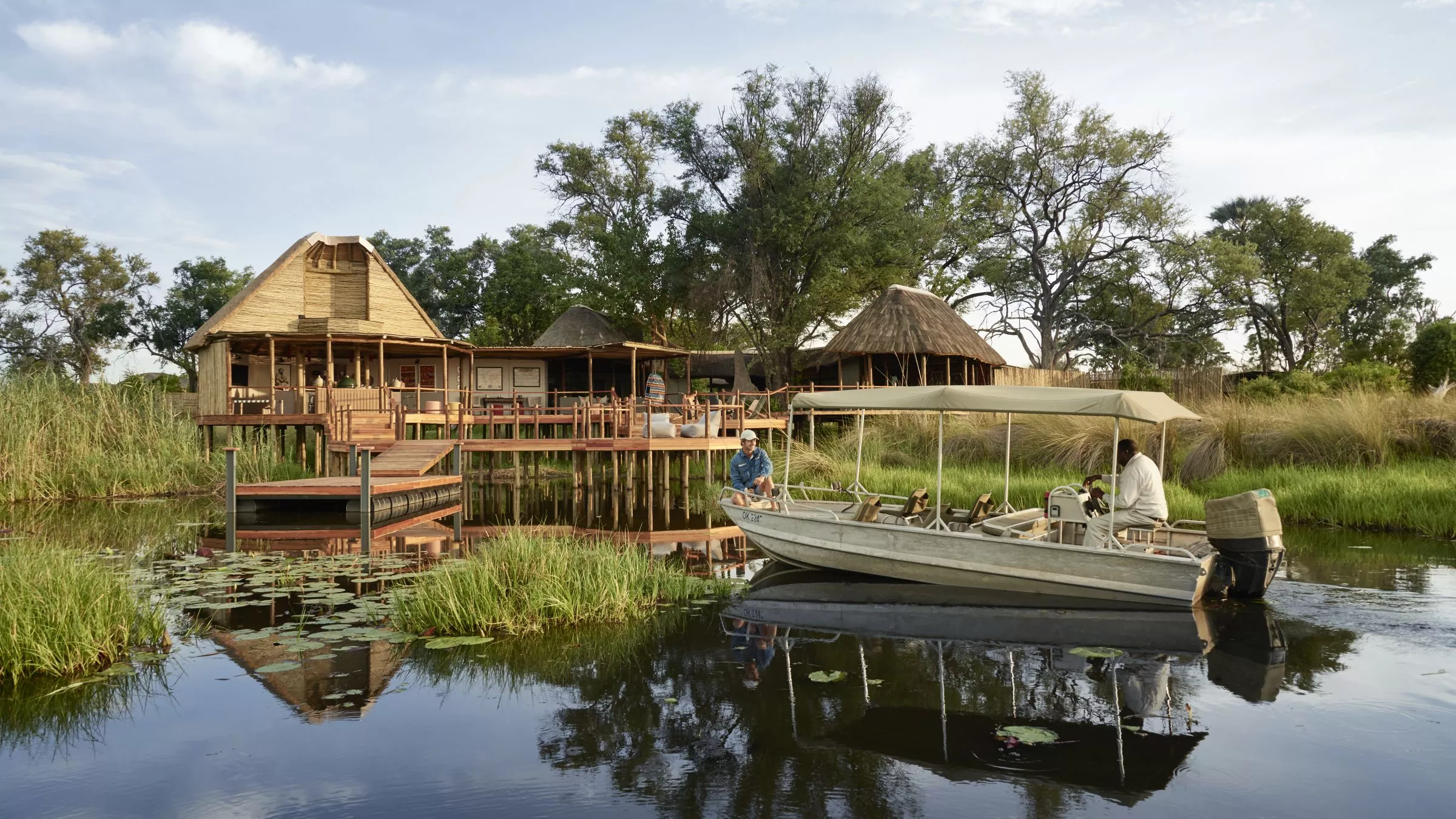 Holzveranda eines Flussresorts mit Strohhäusern; Boot mit Passagieren am Ufer, Wasserreflexionen.