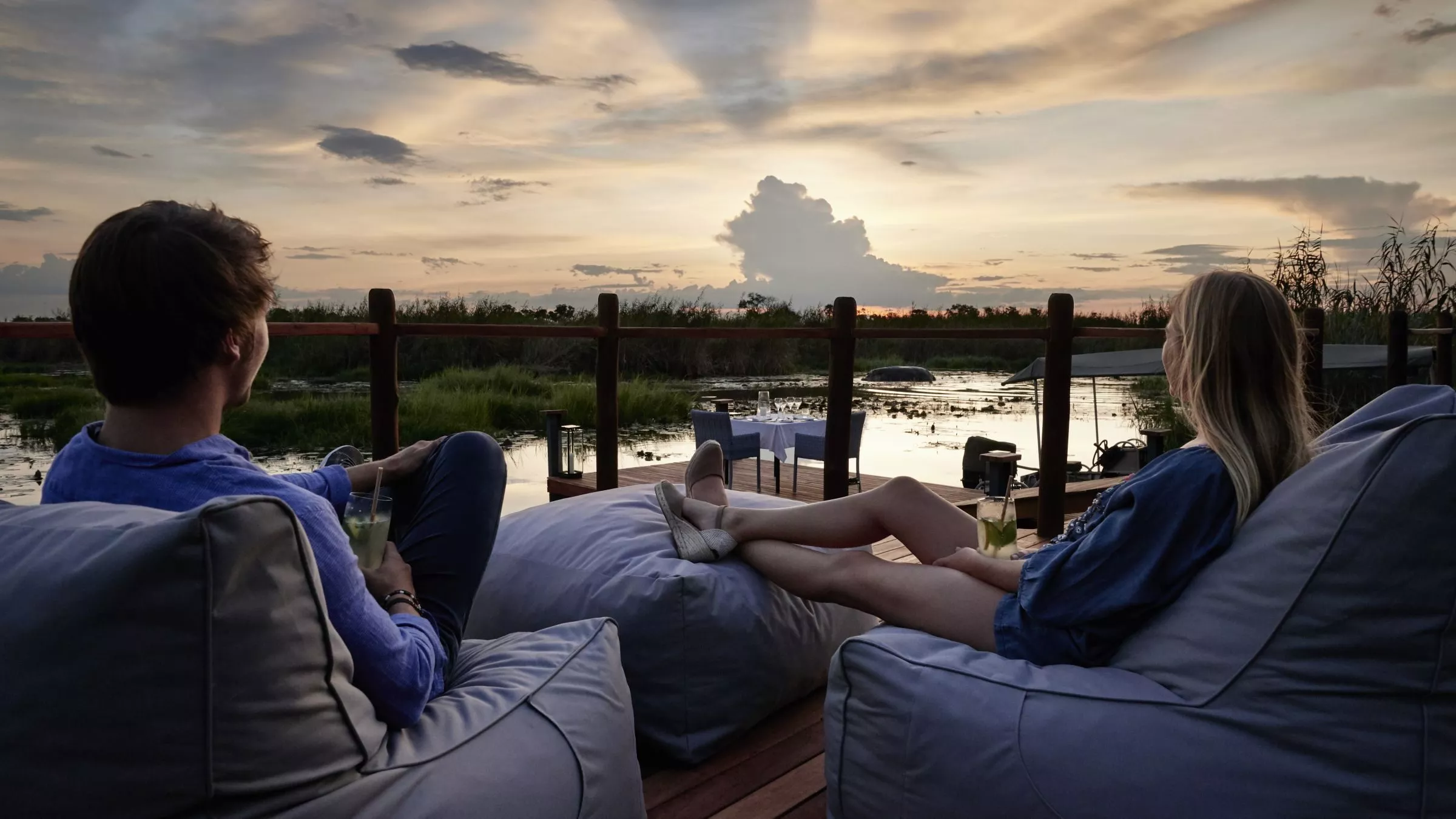 Zwei Personen ruhen auf großen Sitzsäcken auf einer Holzterrasse am Wasser; Sonnenuntergang, Getränke in den Händen.