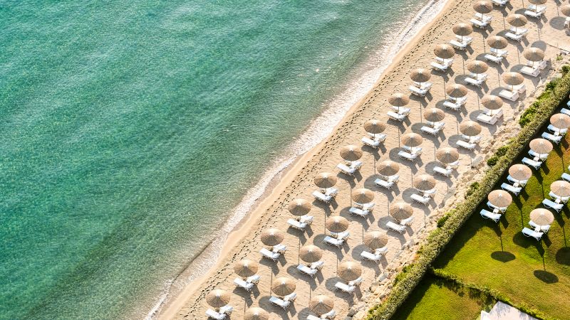 Luftaufnahme eines Strandes mit Reihen von Sonnenschirmen und Liegen am türkisblauen Meer, Domes Noruz Kassandra.