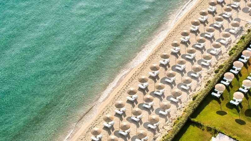 Luftaufnahme eines Strandes mit Reihen von Sonnenschirmen und Liegen am türkisblauen Meer, Domes Noruz Kassandra.