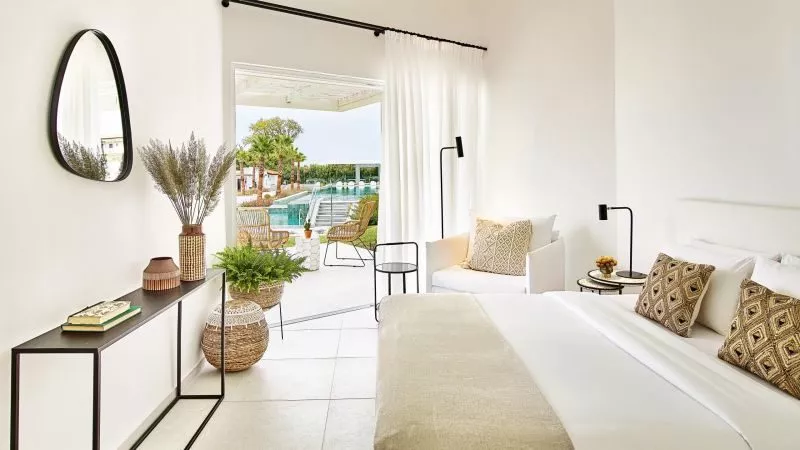 Helles Domes Noruz Kassandra Hotelzimmer: Bett mit beigeem Überwurf, gemusterte Kissen, Konsole mit Pflanzen, runder Spiegel, Tür zum Poolgarten.