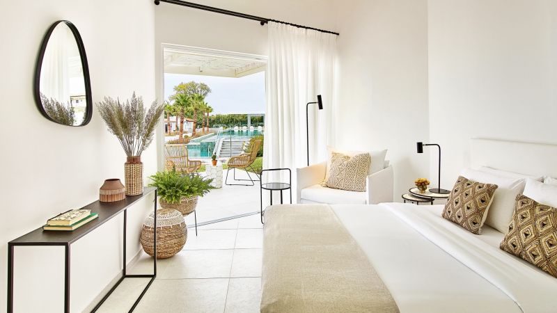 Helles Domes Noruz Kassandra Hotelzimmer: Bett mit beigeem Überwurf, gemusterte Kissen, Konsole mit Pflanzen, runder Spiegel, Tür zum Poolgarten.