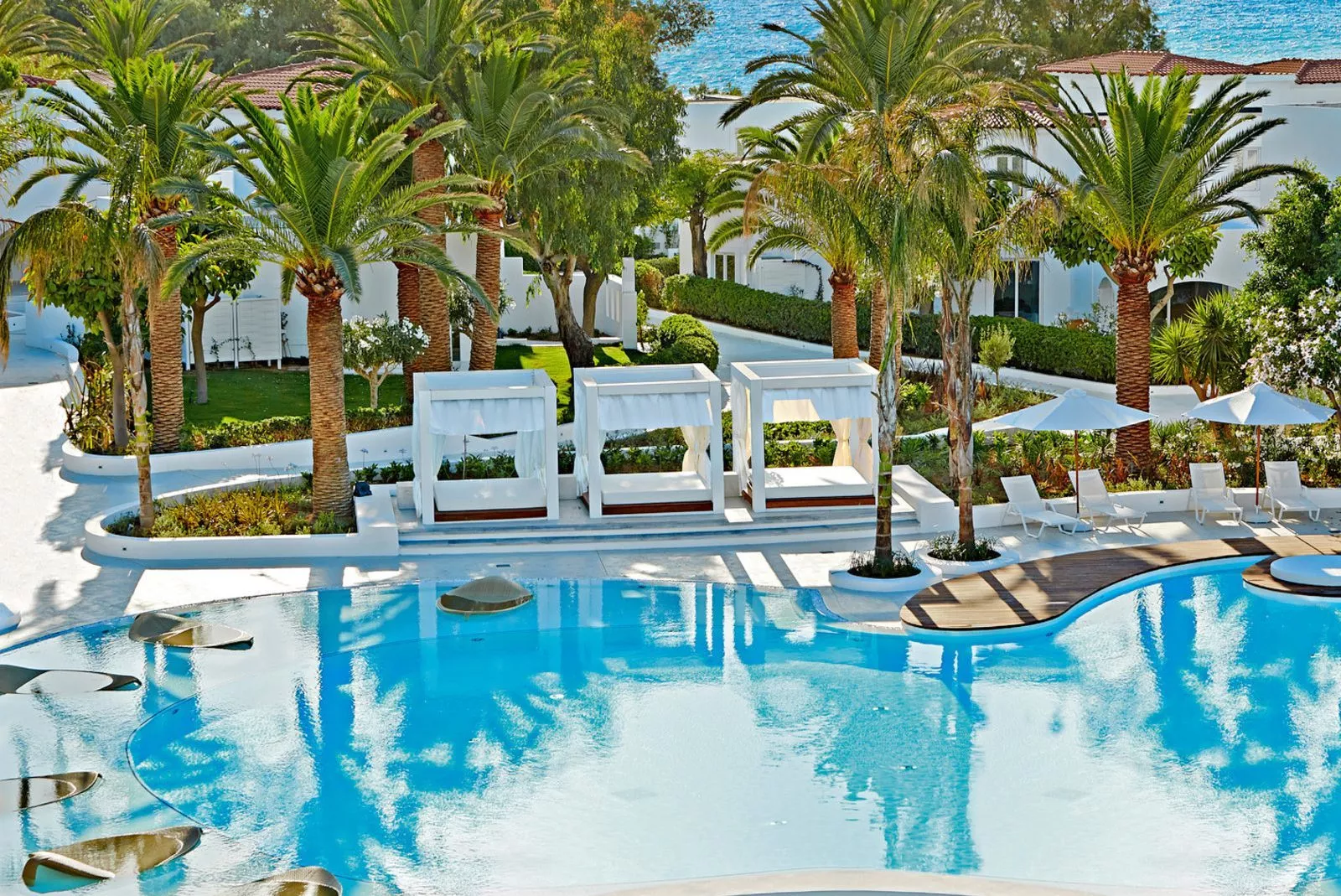 Weißer Poolbereich mit Palmen, drei Cabana-Suiten, Liegen und Sonnenschirmen; Meer dahinter.