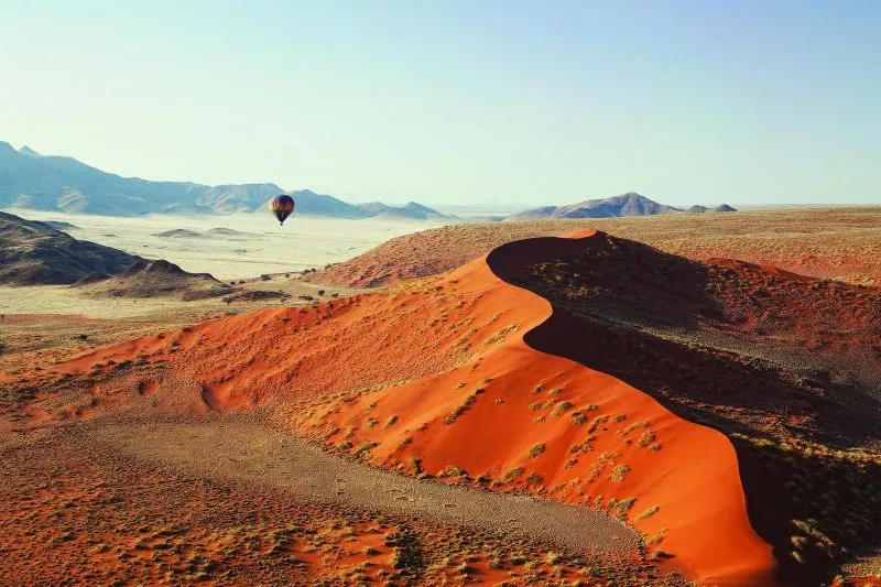 Namibias Wüstenlandschaft: rote Dünen im Vordergrund, weite Ebene und Berge; Heißluftballon am Horizont.