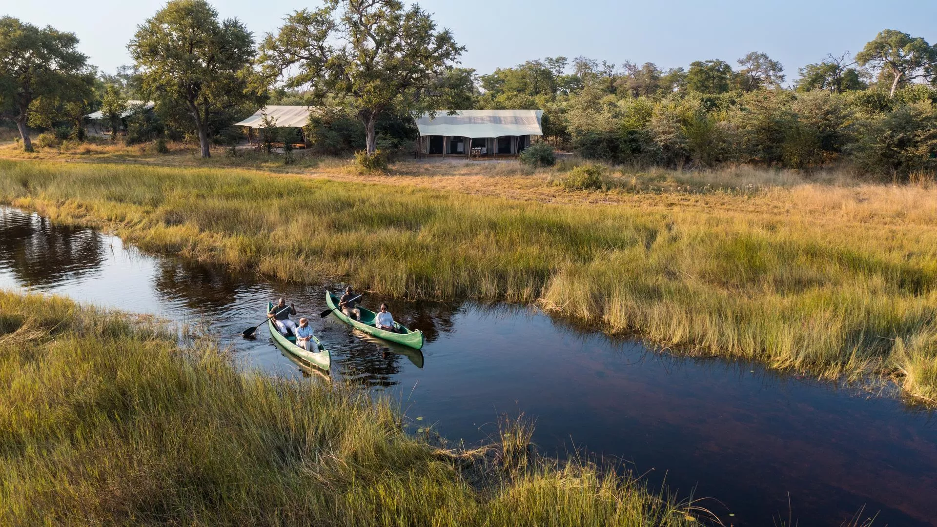 Flusslandschaft mit grasbewachsenem Ufer; drei grüne Kanus mit Paddlern, dahinter Zeltlager unter Bäumen in Botswana
