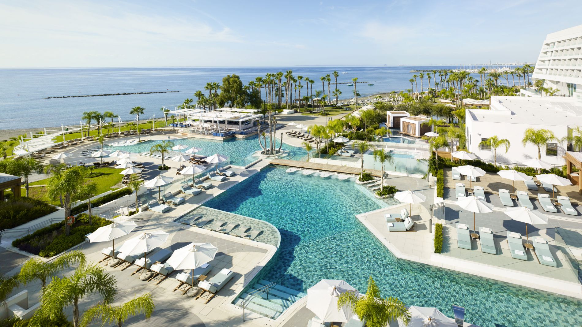 Luftaufnahme des Parklane Limassol Resorts am Meer: mehrere Poolbereiche, Palmen, Liegen mit Sonnenschirmen, Strandpromenade.