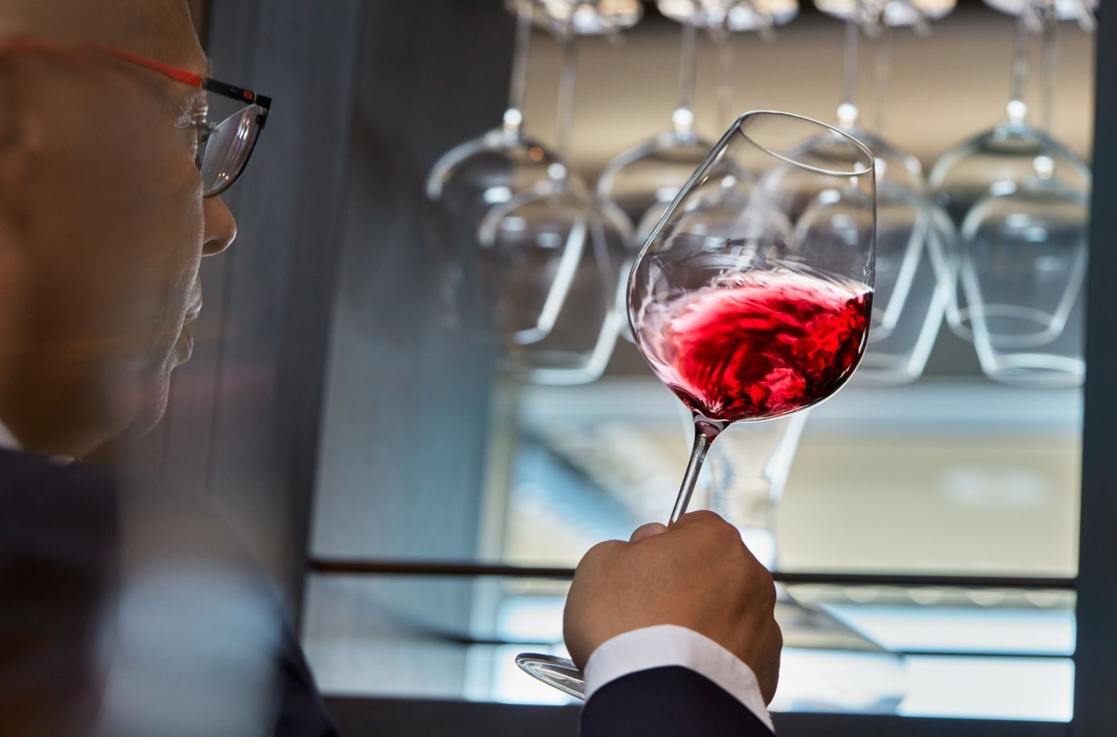 Mann mit Brille betrachtet ein Rotwein-Glas in Nahaufnahme; Hintergrund Gläserregal.