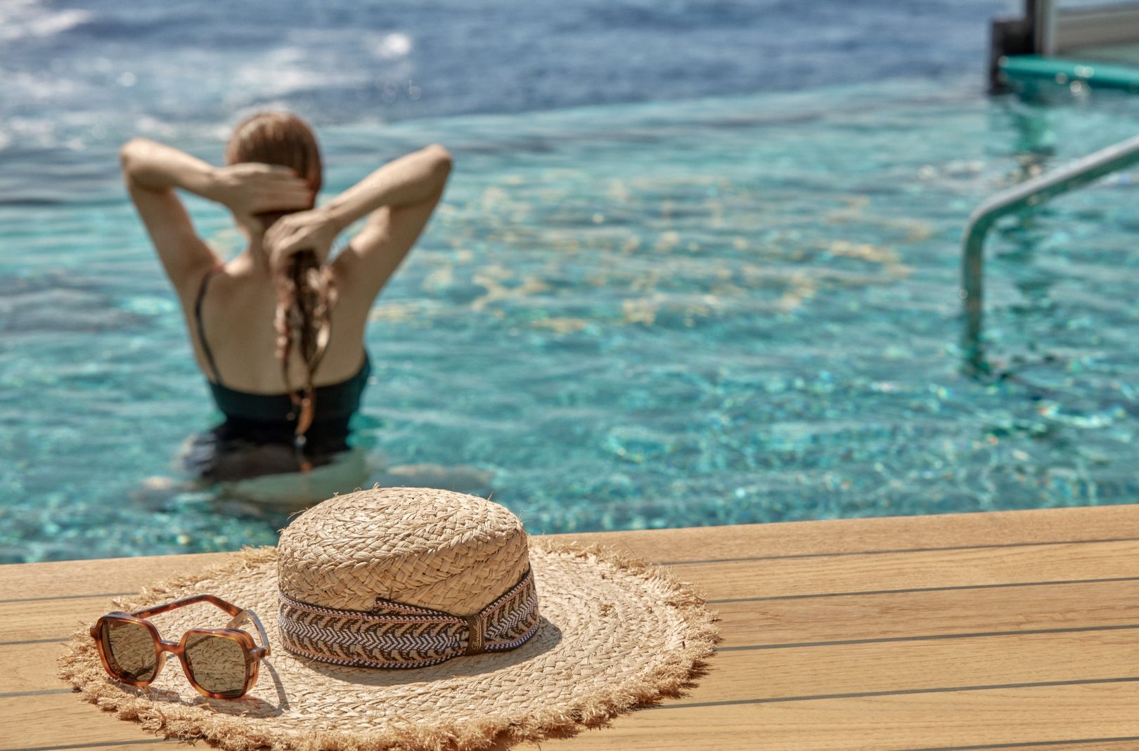 Frau im Pool von Explora Journeys, Rückenansicht beim Entspannen; Strohhut und Sonnenbrille liegen auf dem Holzdeck.