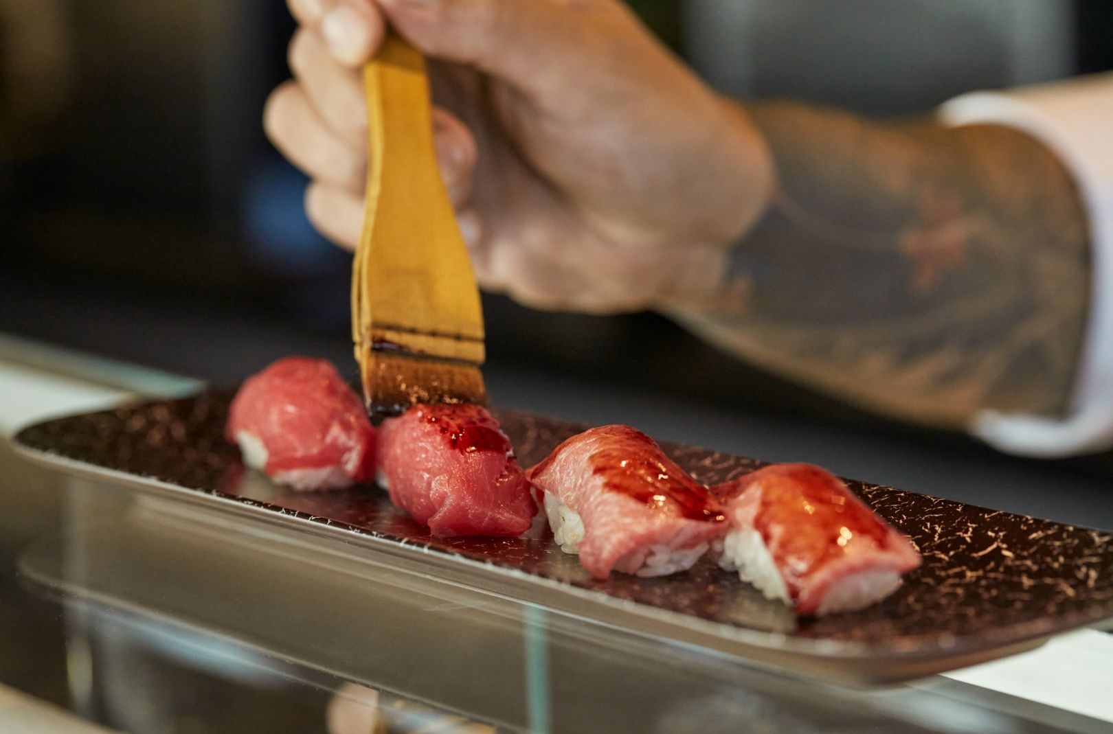Koch bestreicht Sushi-Nigiri auf dunklem Teller mit Pinsel; tätowierter Arm im Hintergrund, Explora Journeys.