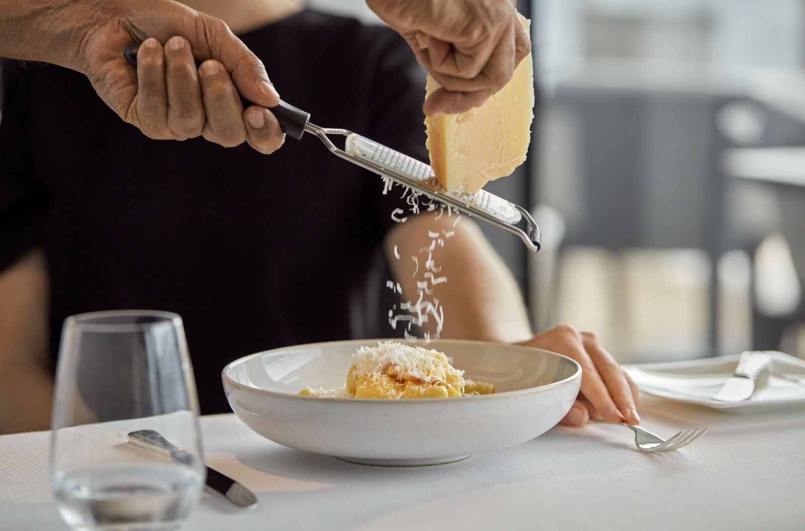 Person reibt Käse über Pasta in einer Schale, Käsehobel in der Hand, Späne fallen; Glas Wasser und Besteck am Tisch.
