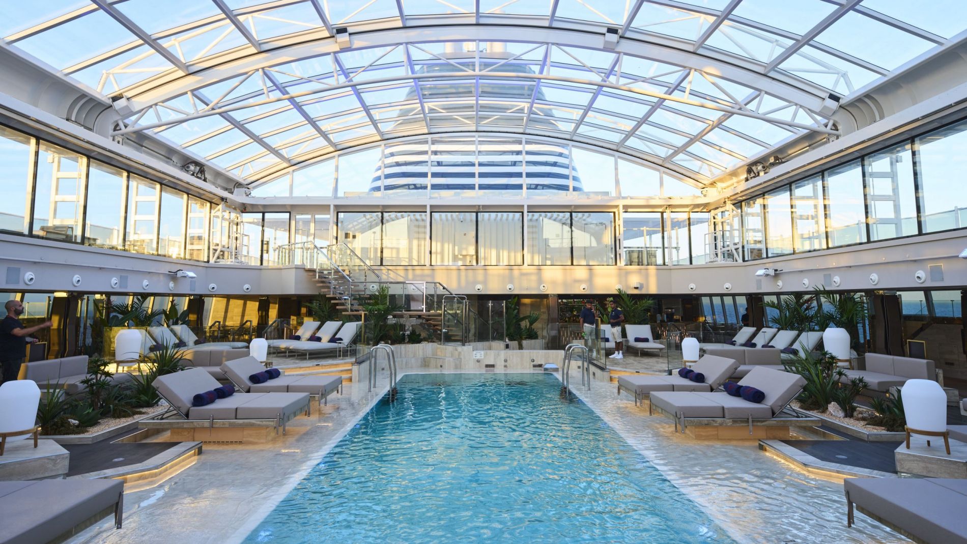 Innenpool unter großem Glasdach, umgeben von Lounge-Liegen, Pflanzen und Glasgalerie im Hintergrund.