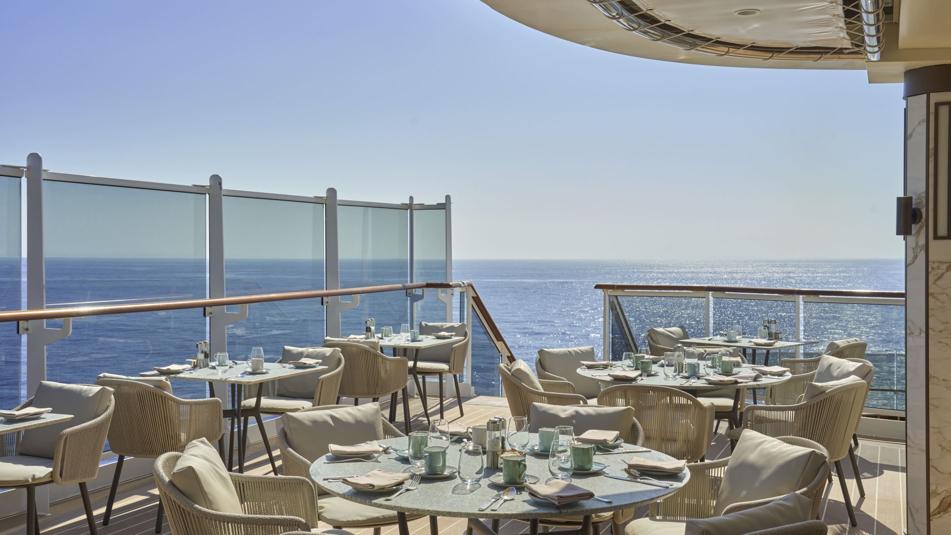 Außenrestaurantdeck mit runden Tischen und Stühlen, Gläsern und Besteck; Glasgeländer, Blick aufs offene Meer.