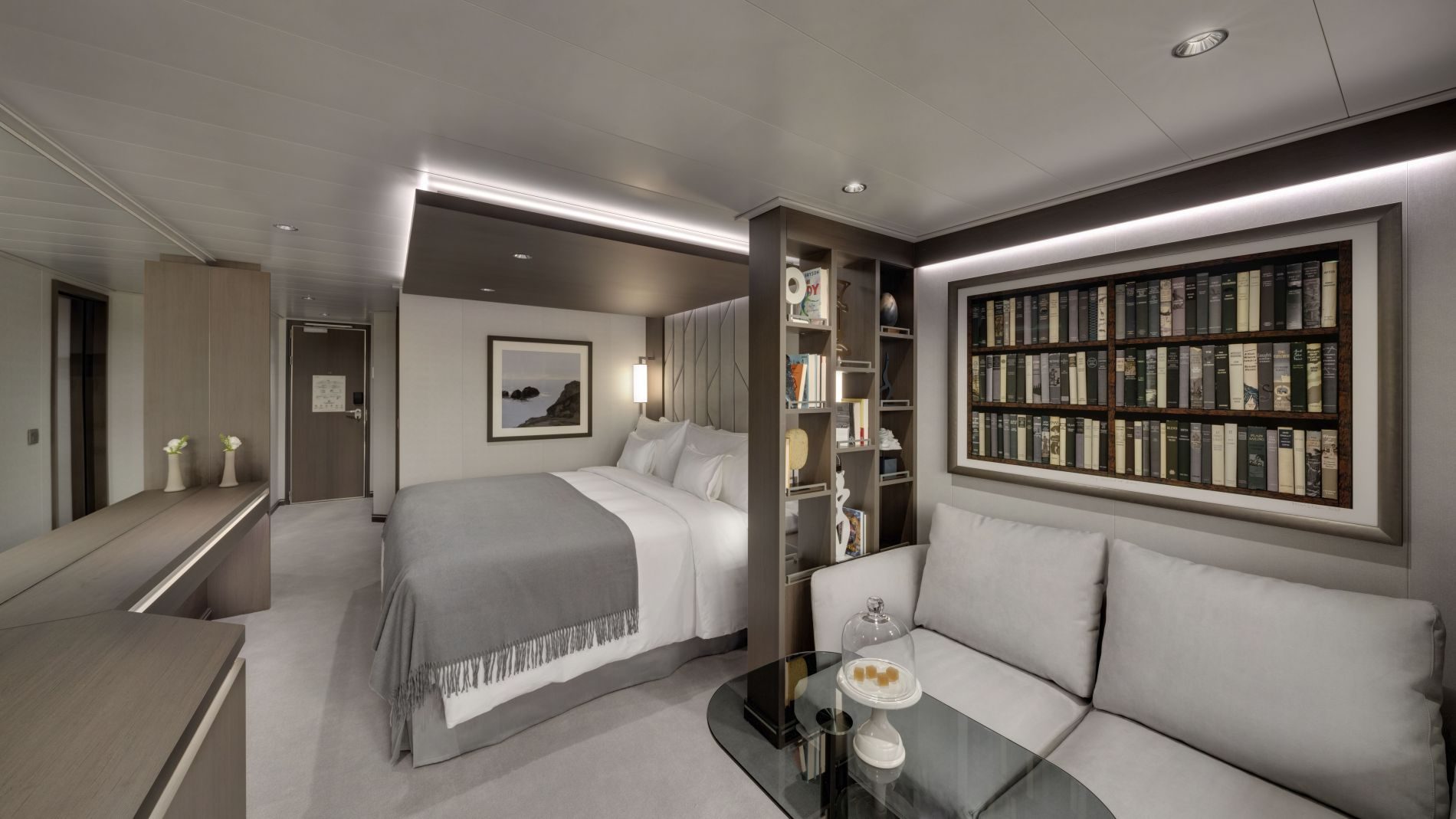 Helles Kabinenzimmer von Explora Journeys: Doppelbett mit grauem Überwurf, Sofa, Glas-Tisch und Bücherregal.