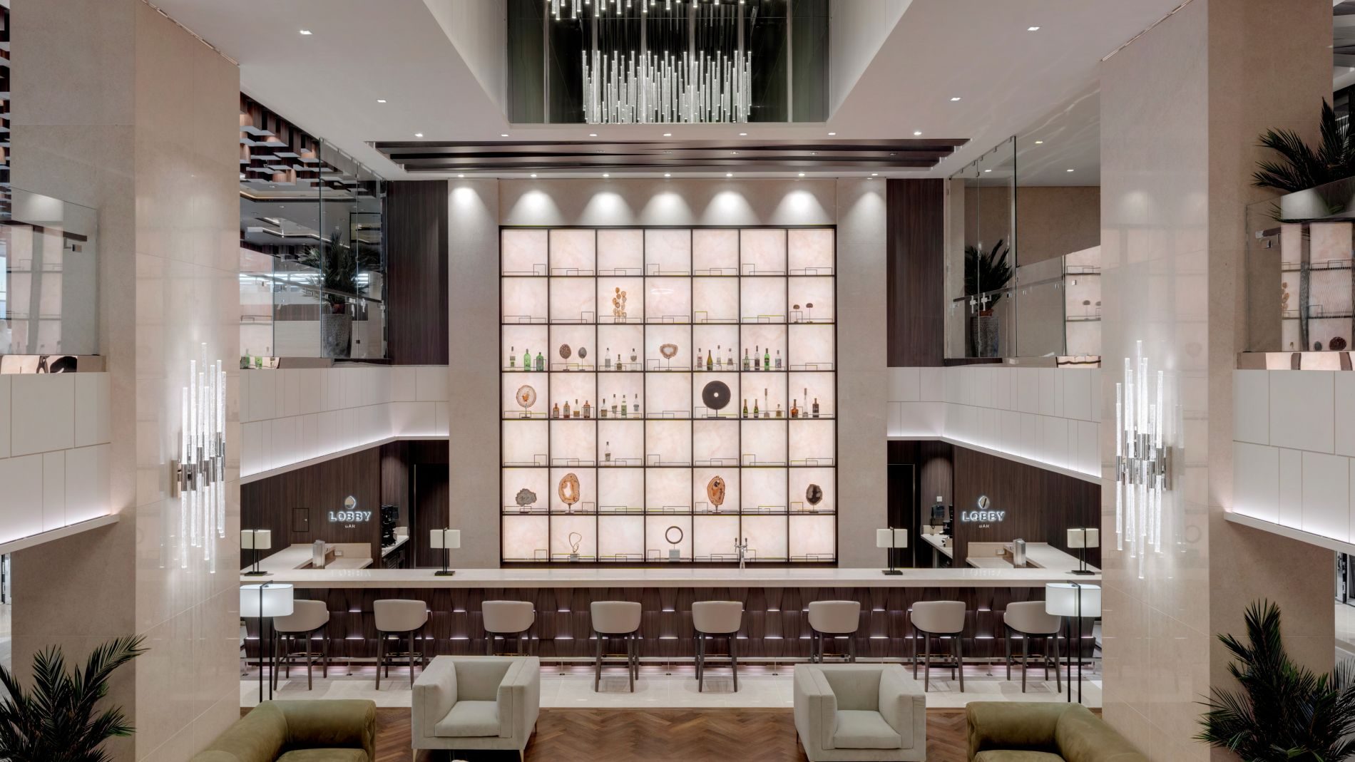 Heller Lobbyausschnitt mit zentraler Bar, beleuchteter Glasregalwand mit Spirituosen, Lounge-Sessel und Sofas