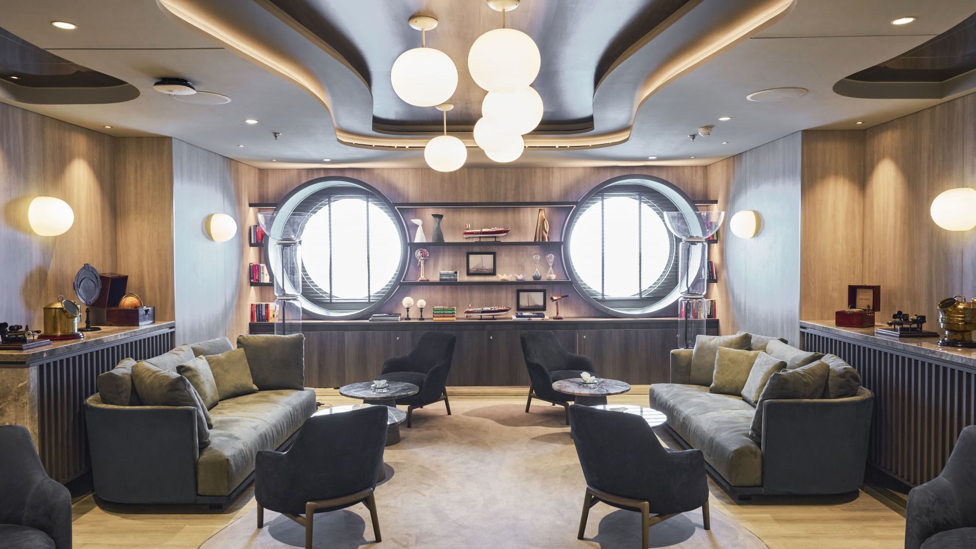 Lounge des Explora Journeys mit zwei runden Fenstern, bequemen Sofas, Sesseln, Couchtischen und Deko-Regalen.
