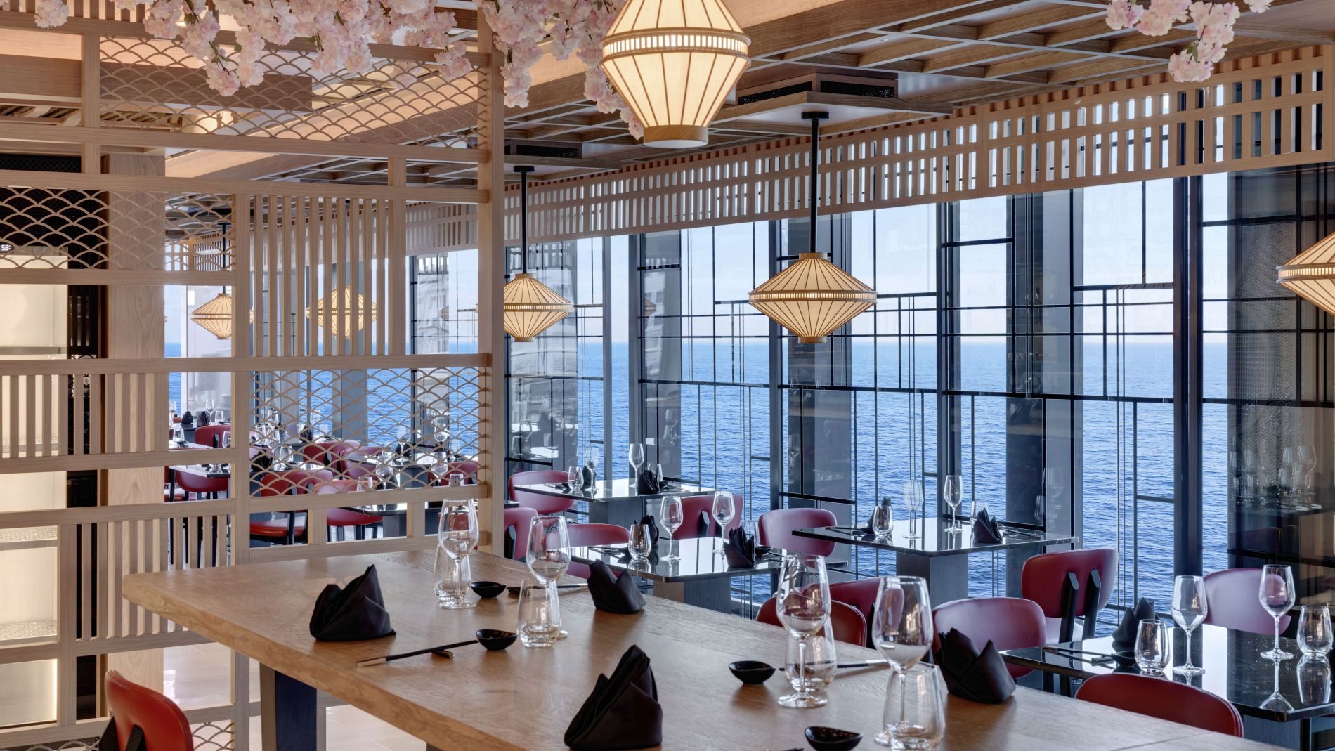 Restaurant mit Holzwandgitter, Kirschblüten-Deko, hängenden Lampen, Meerblick durch Fenster, gedeckte Tische mit Gläsern.