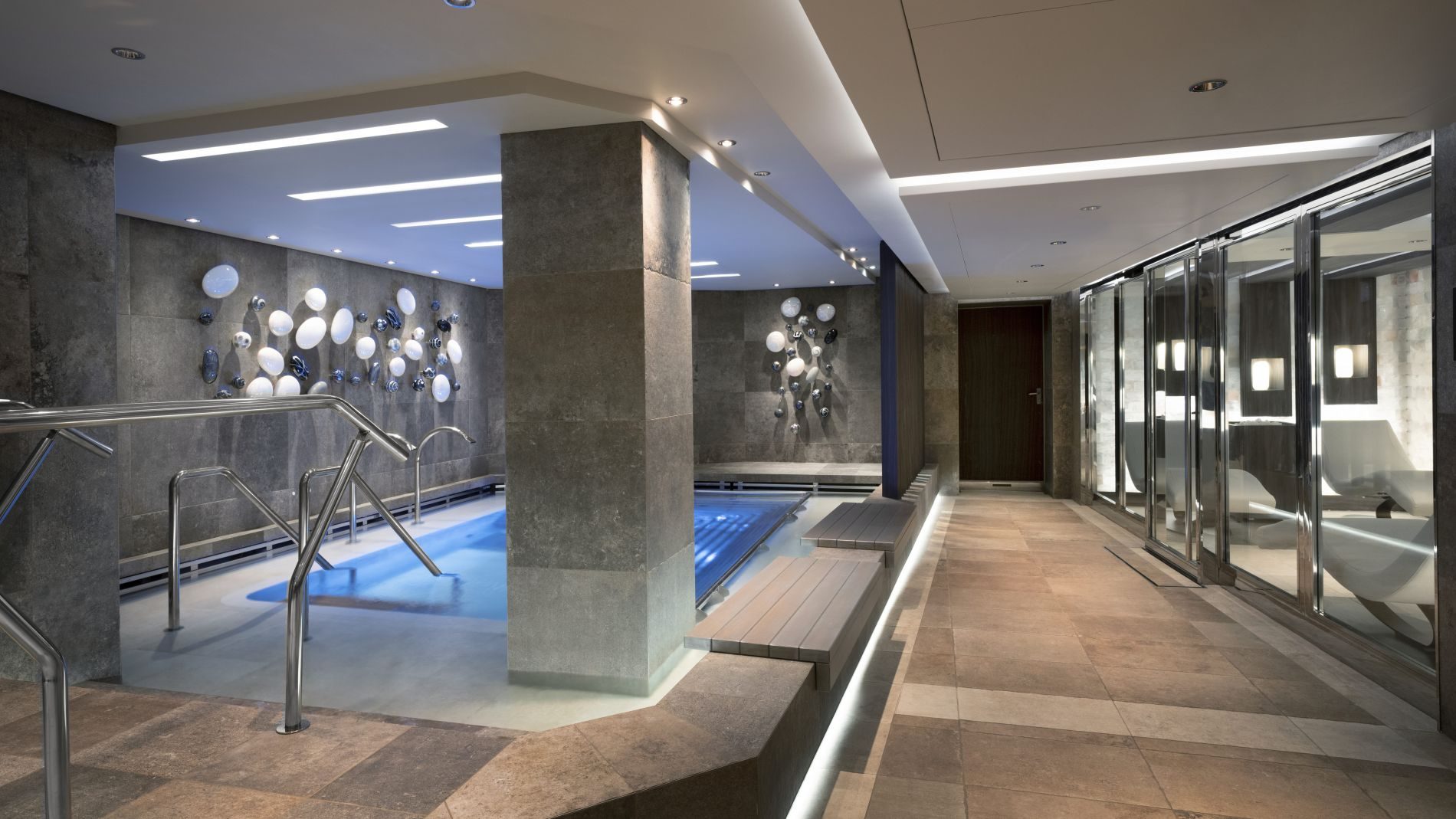 Wellnessbereich mit kleinem Becken, Metallhandläufen, Steinfliesen und kreisförmiger Wandkunst; Glasfronten rechts
