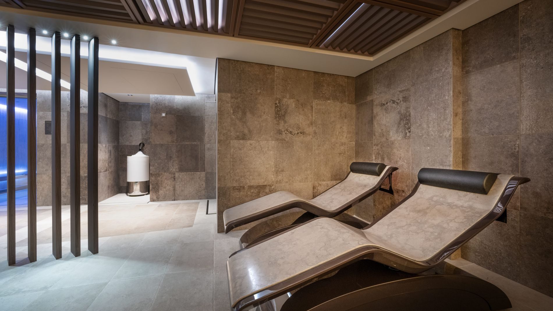 Spa-Lounge mit zwei geneigten Steinliegeflächen, dunkle Steinwand, Holzdecke, vertikale Trennstangen; links blauer Beckenbereich.