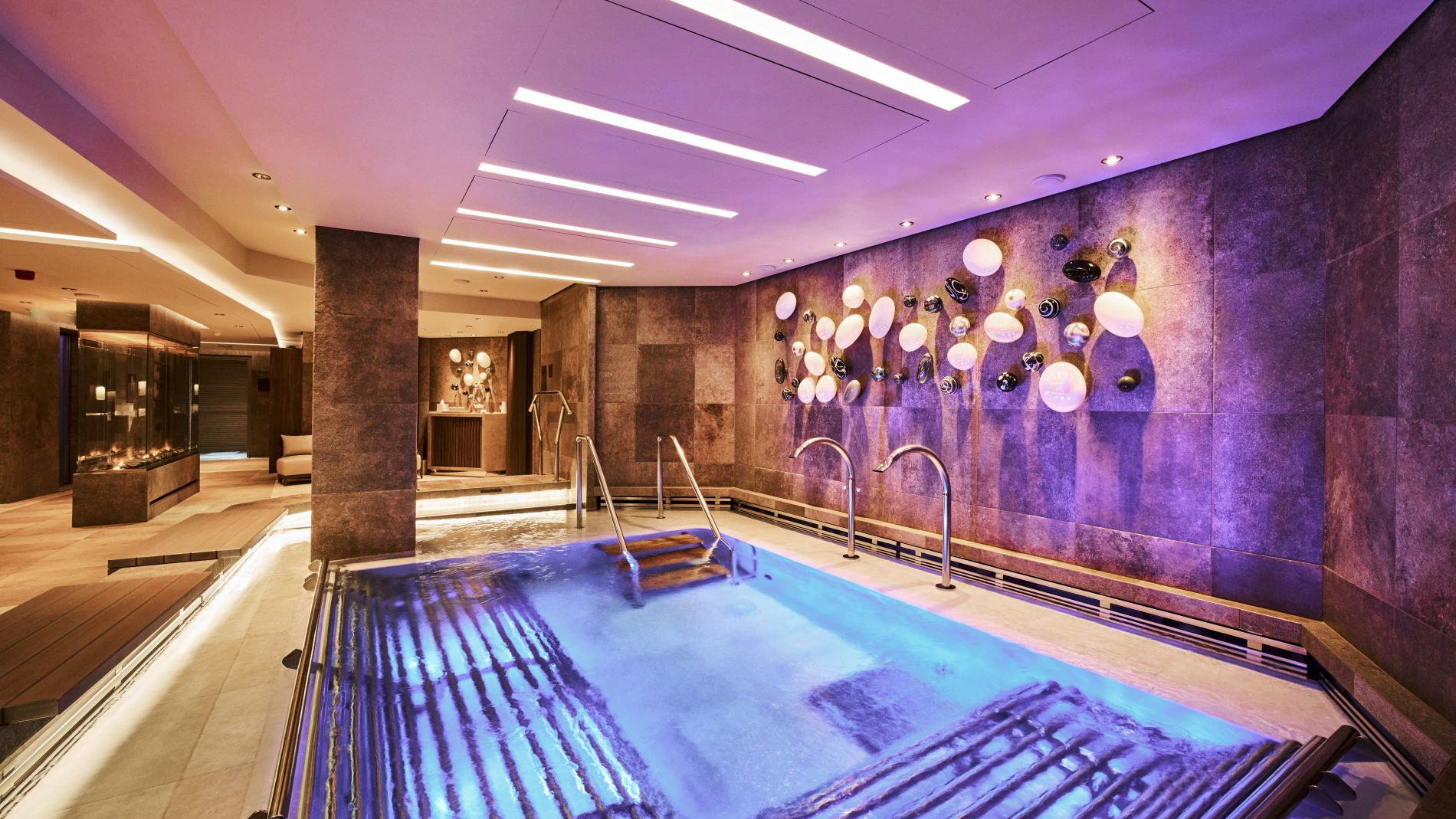 Spa-Poolbereich im Wellnesslayout von Explora Journeys; bläulich-lila beleuchtetes Wasser, Treppenstufen, Metallhandläufe und Wandkunst aus Kugeln.