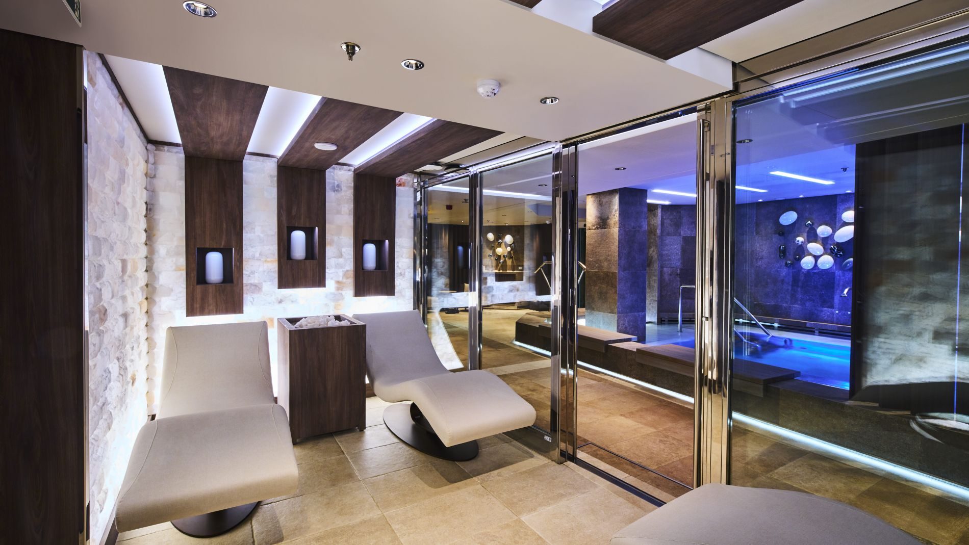 Wellness-Lounge mit Holzpaneelen, Steinwand, grauen Lounge-Sesseln und Glasfronten zum Poolbereich Explora Journeys.