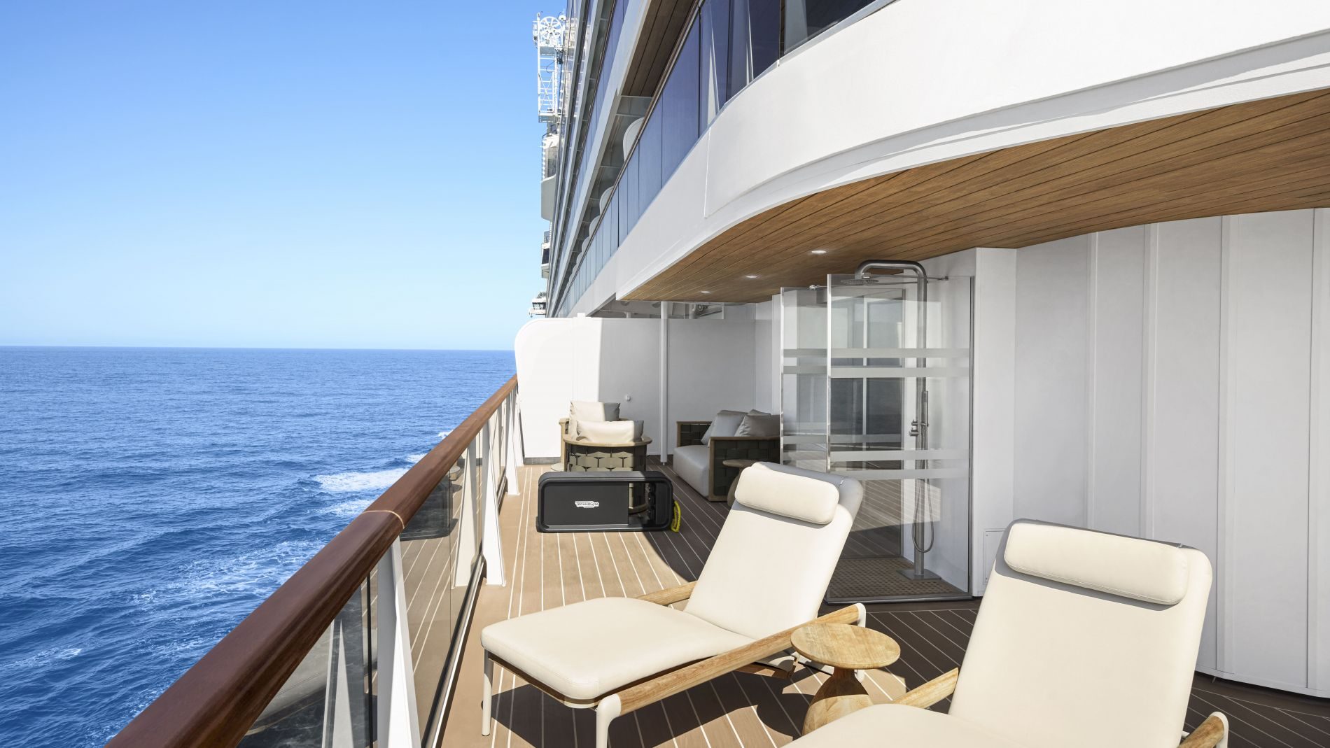 Außendeck eines Explora Journeys Schiffes: beige Lounge-Sessel, Beistelltisch, Glasgeländer, Holzdeck, Blick aufs Meer.