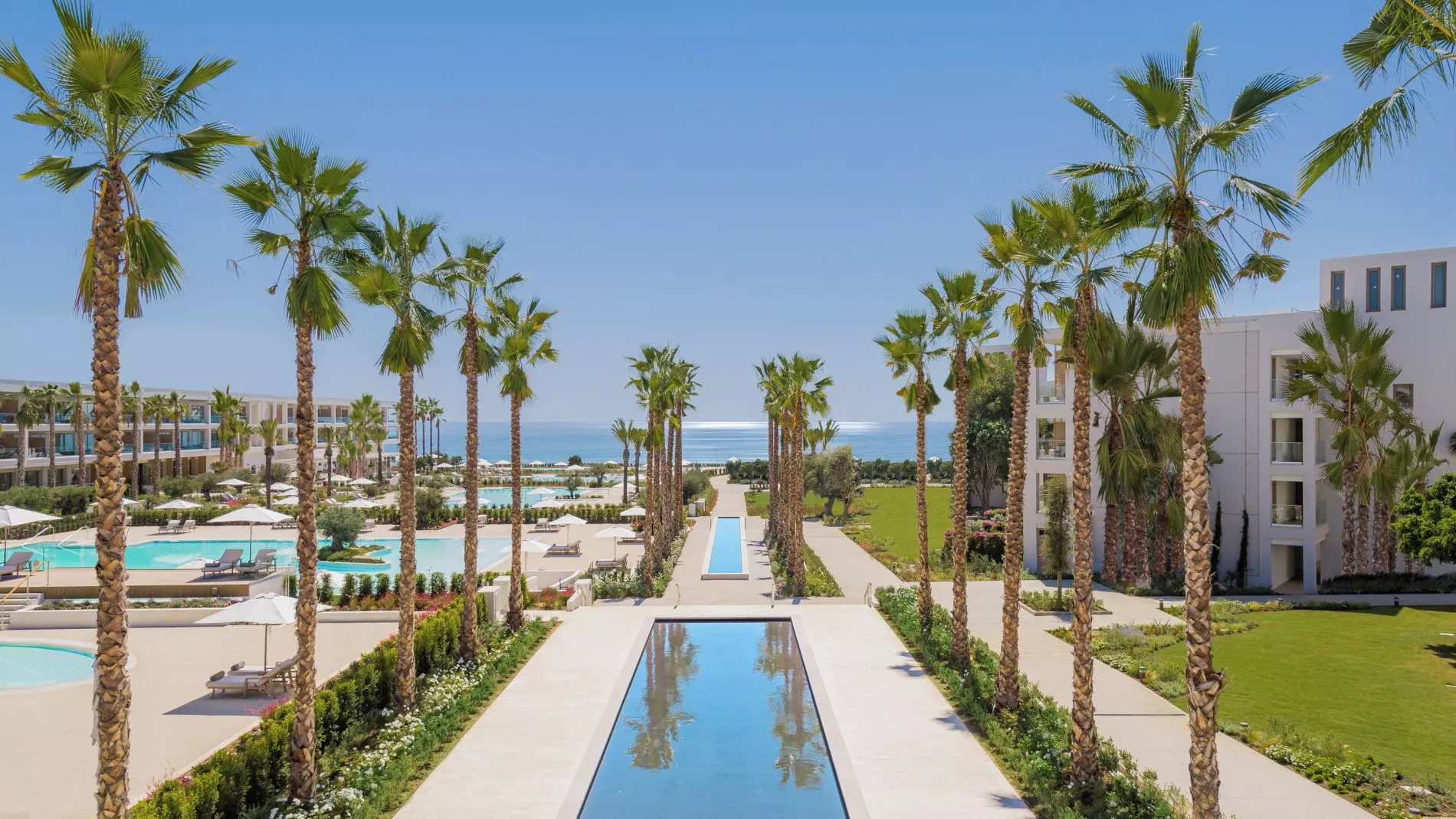 Resortansicht: Palmen säumen einen zentralen Wasserkanal, Poolbereich mit Liegen, weiße Hotelgebäude, Blick aufs Meer.