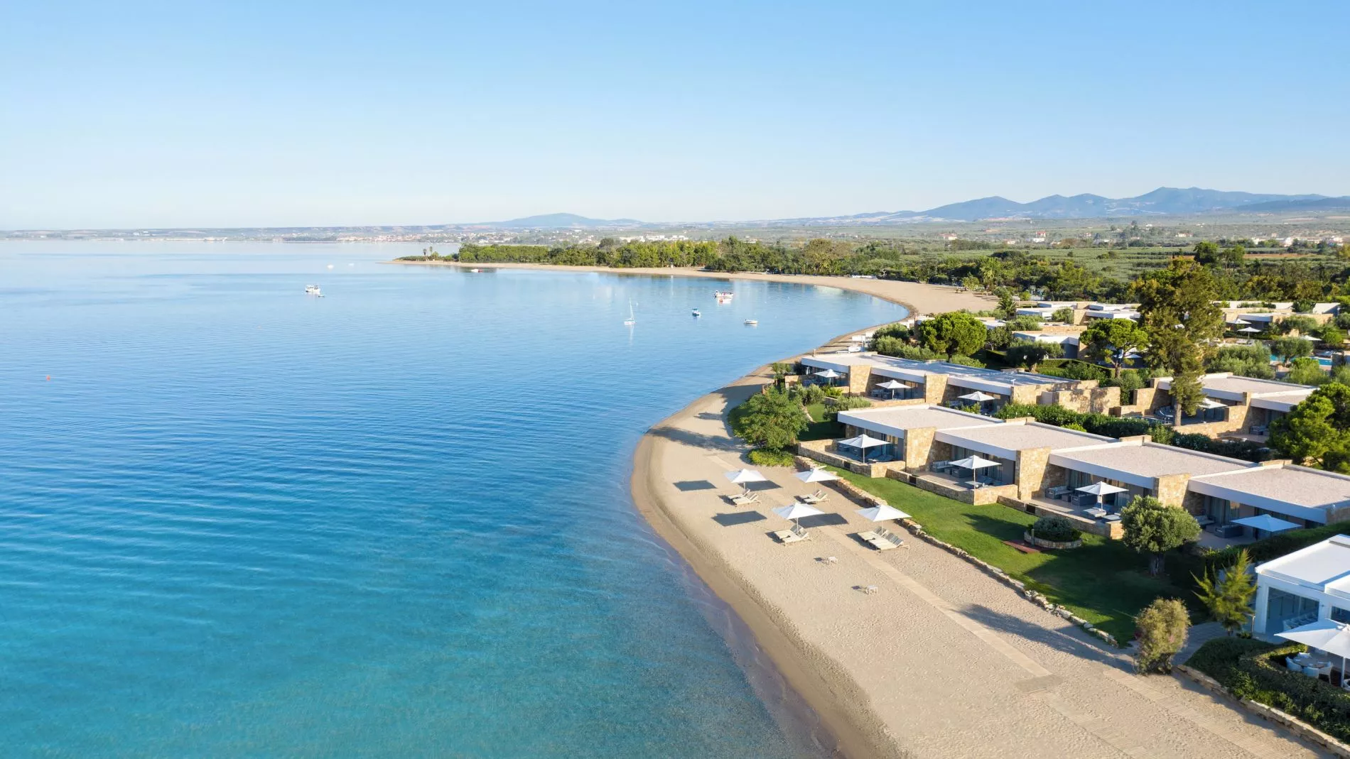 Luftaufnahme der Ikos Olivia Deluxe Beach Area: Sandstrand, türkisblaues Meer und Reihen von Beachvillen mit Sonnenschirmen.
