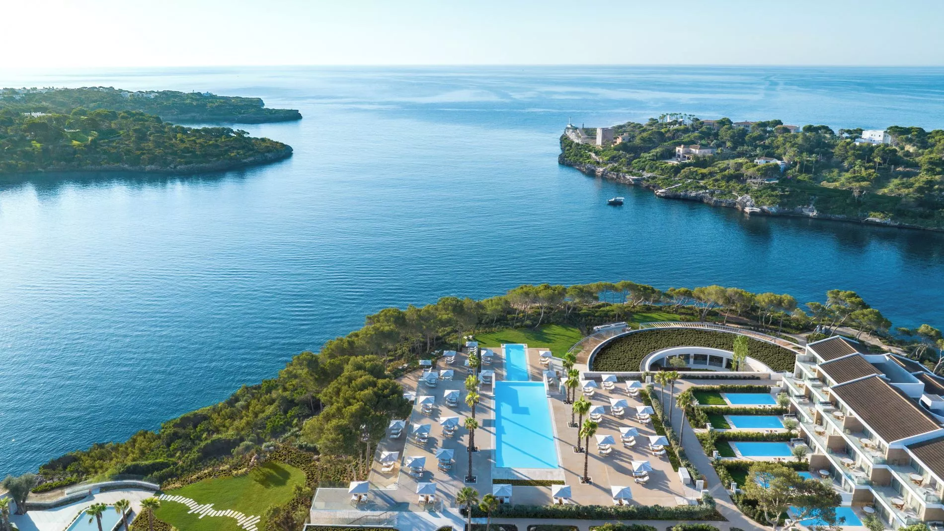 Luftbild eines Hotelresorts am Meer mit langem Pool, Liegen, Palmen und Gartenlandschaft, Blick aufs offene Wasser.