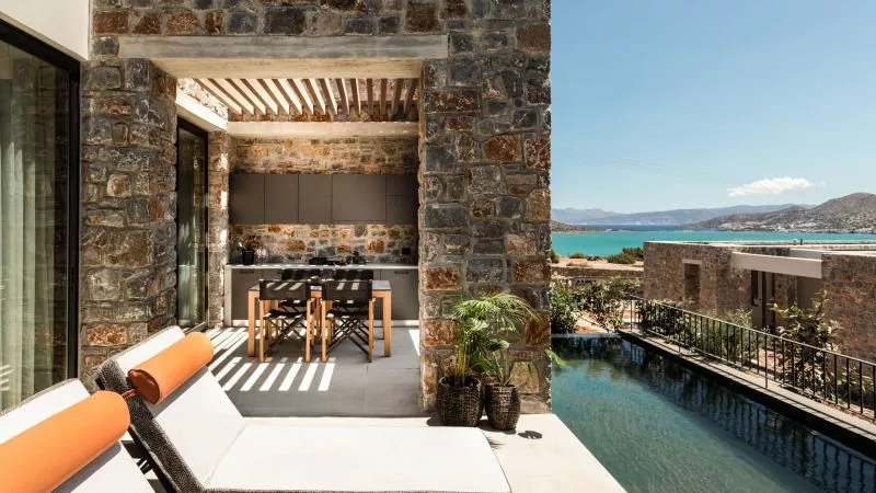 Terrasse aus Stein mit Lounge, Esstisch und Pool; Blick aufs Meer und die Hügel von Elounda.