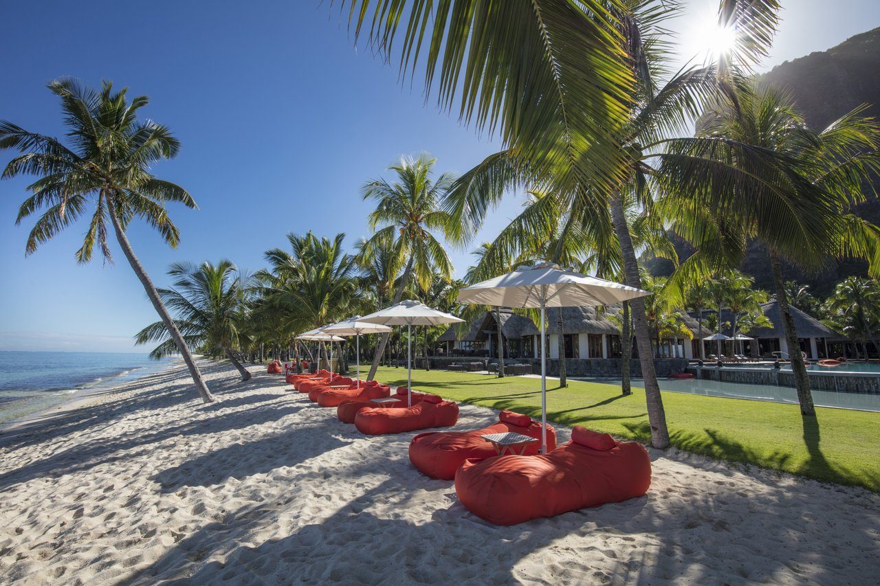 Strand mit Palmen, rote Beanbags unter Sonnenschirmen, Blick aufs Resort Dinarobin Beachcomber am Meer. Strand mit Palmen, rote Beanbags unter Sonnenschirmen, Blick aufs Resort Dinarobin Beachcomber am Meer.