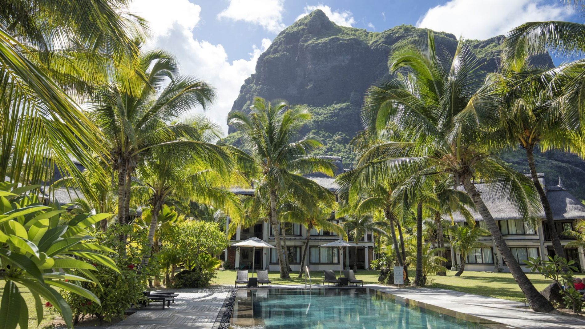 Poolbereich mit Liegen und Palmen, Hotelgebäude im Garten, Berglandschaft im Hintergrund, Beachcomber Resorts & Hotels. Poolbereich mit Liegen und Palmen, Hotelgebäude im Garten, Berglandschaft im Hintergrund, Beachcomber Resorts & Hotels.