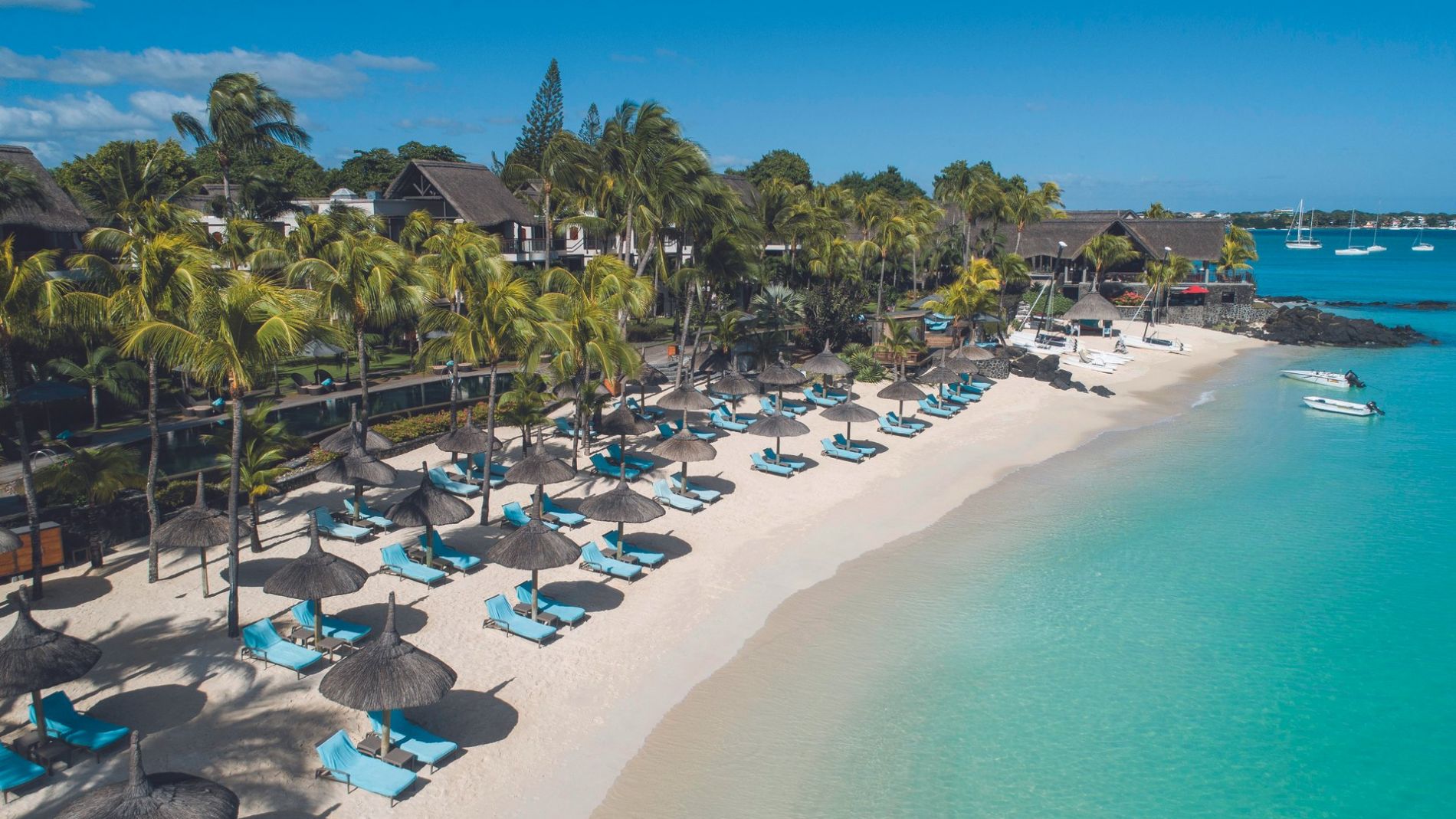 Strand mit Palmen, Strohdach-Schirmen und blauen Liegen am klar türkisfarbenen Meer; Hotelanlage im Hintergrund. Strand mit Palmen, Strohdach-Schirmen und blauen Liegen am klar türkisfarbenen Meer; Hotelanlage im Hintergrund.
