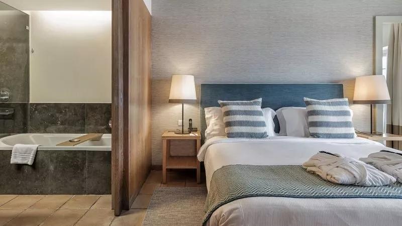 Hotelzimmer mit Doppelbett, blauem Kopfteile, zwei Nachttische mit Lampen, gestreifte Kissen, Badewannenbereich hinter Holztrennwand sichtbar