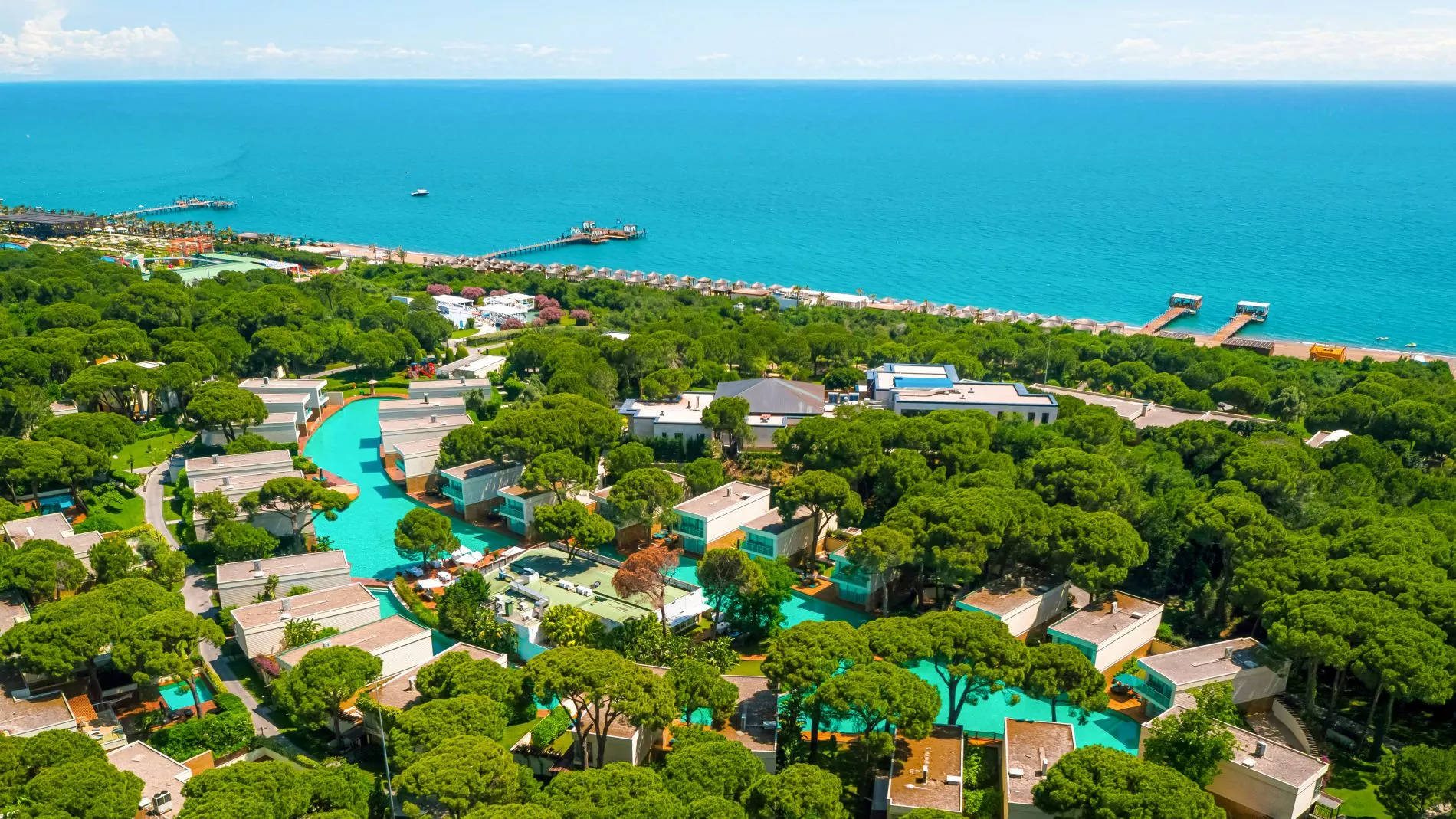 Luftaufnahme eines Resortkomplexes in Belek am Meer mit grünem Baumbestand, vielen Pools und Strandpromenade.