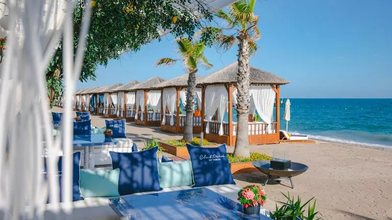 Strandlounge mit Holzcabanas und weißen Vorhängen, Palmen, blaue Kissen, Tischplatte im Vordergrund, Blick aufs Meer.