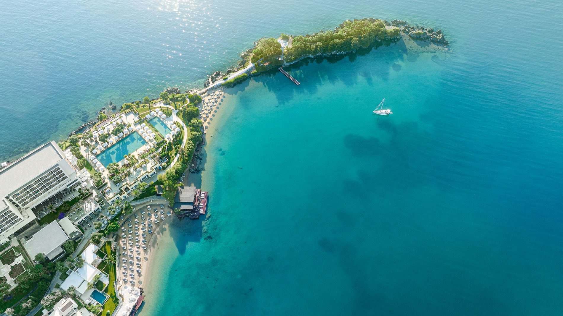Luftaufnahme des Corfu Imperial Resorts mit Pool, Strandabschnitt und Boot vor einer Halbinsel im türkisblauen Meer.